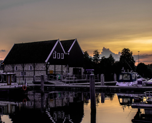 Boten aangemeerd in een jachthaven bij zonsondergang, met een traditioneel gebouw op de achtergrond.
