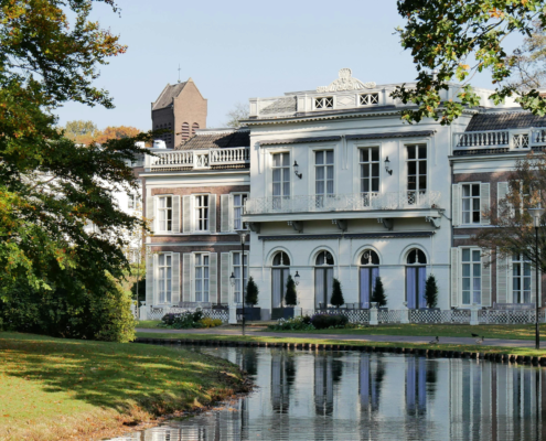 Elegante villa met witte luiken aan een rustige vijver, omgeven door bomen in herfstkleuren.