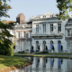 Elegante villa met witte luiken aan een rustige vijver, omgeven door bomen in herfstkleuren.